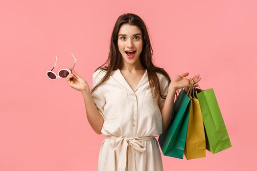 Cheerful gorgeous caucasian woman in dress, holding shopping bags and sunglasses, smiling intrigued and excited, seeing beautiful garment on stalls want buy, standing pink background.