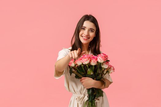 You deserve best. Charming lovely and romantic, tender caucasian girl in dress, holding roses near chest, wrap bouquet gently and pointing finger camera, smiling delighted, standing pink background.