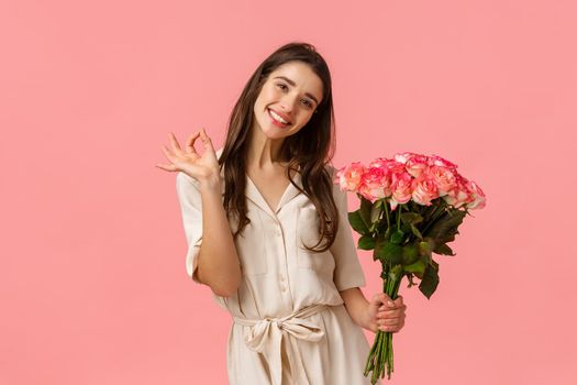 Lovely, dreamy and coquettish brunette female in dress, tilt head and smiling delighted, showing okay, good holding roses, got gift on valentines day, beautiful bouquet flowers, pink background.