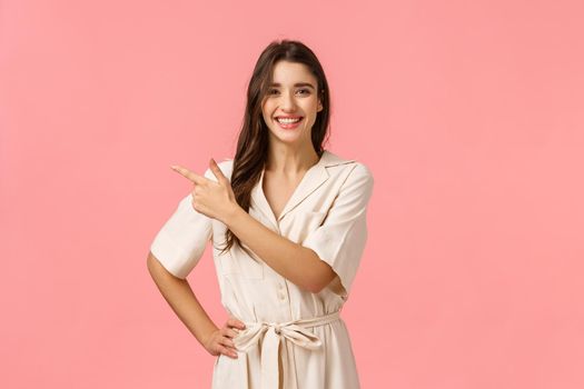 Enthusiastic positive cute brunette girl in pretty dress, pointing upper left corner, gladly showing product discounts promo, standing pink background, smiling joyfuly and excited.