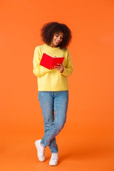 Full-length vertical shot dreamy cute african-american female student with afro haircut, casual clothes, going college, making notes, standing over orange background and writing in notebook smiling.