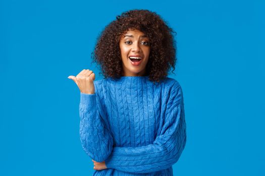 Excited happy pretty african-american woman with afro haircut in winter sweater, pointing thumb left and laughing as discuss funny moment, talking casually during party, blue background.