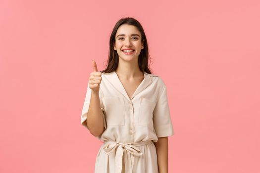 Giving positive feedback, recommendation. Good-looking cheerful, tender girl in gorgeous dress, showing thumb-up in approval, satisfied or advertising gesture, smiling like product, pink background.