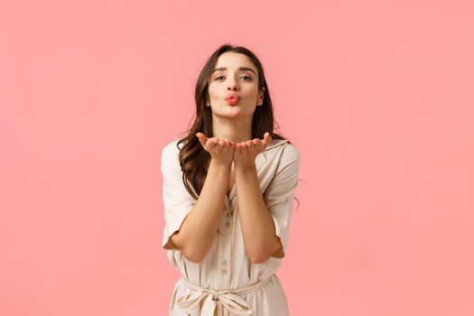 Sending love and care. Charming cute and tender european brunette with curly hair, wearing spring dress, leaning with hands near folded lips, blowing air kiss with love, standing pink background.