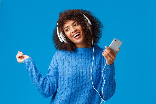 Joyful and carefree lovely african-american female model in winter sweater, dancing to rhythm music, listening new track on song app, christmas playlist, wear headphones hold smartphone.