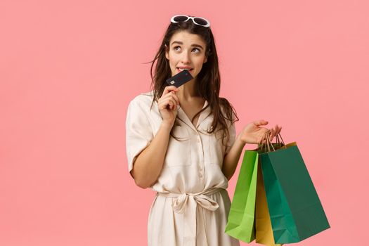 Girl eager waste more money feeling guilty biting credit card and looking thoughtful hesitant up, thinking, trying stop buying new things, shoppaholic holding shopping bags, pink background.