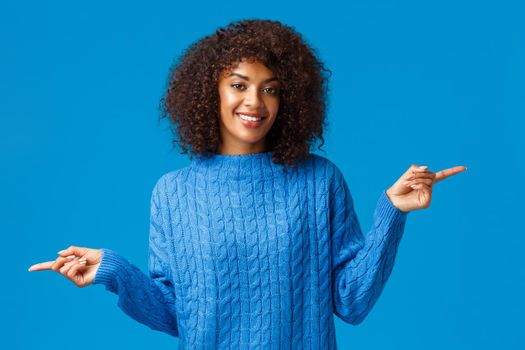 Both are good, take a look. Pleasant gorgeous african-american girl in winter sweater, suggest two variants, pointing fingers sideways, left and right promos, smiling cheerful, blue background.