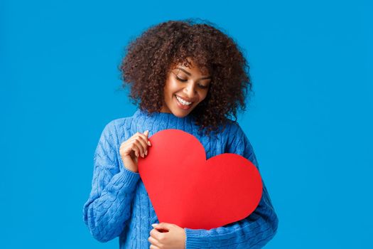 Tenderness, sympathy and relationship concept. Dreamy lovely african-american girlfriend in love, seeking soulmate, found her couple, holding red big heart sign and smiling silly, blue background.