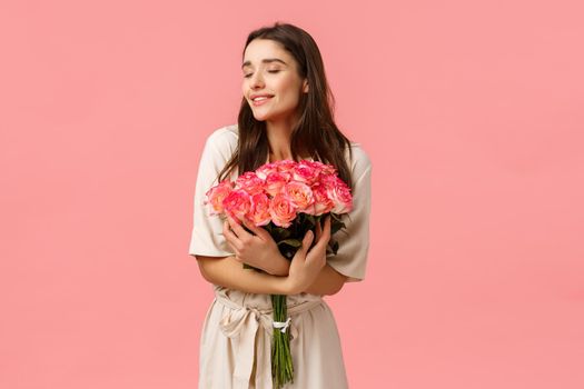 Tenderness, beauty and romance concept. Romantic good-looking tender brunette girl in dress, smiling sensually and delighted, holding roses, receive bouquet roses and laughing, pink background.