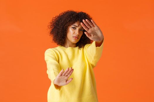 No stop, turn-off lights. Displeased grumpy cute african-american woman with afro haircut cover face from spotlight, defending from glimmering light, standing orange background reluctant.