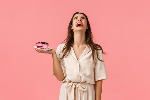 Sad and heartbroken lonely young girl broke-up with boyfriend crying and sobbing, holding cake, eating to ease pain, standing sulky and unhappy over pink background, express sadness and distress.
