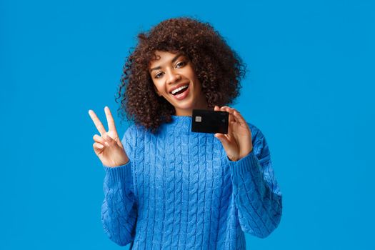 Cheerful cute african-american woman with afro haircut, tilt head show peace sign and holding credit card, got everything for holidays purchase online, standing blue background joyful.