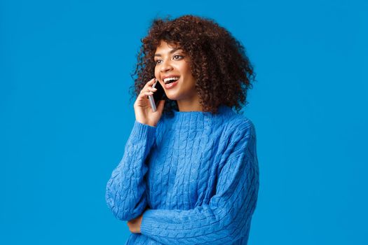 Girl congratulating family with happy holidays, wishing well new year as talking on phone from abroad, holding smartphone speaking, calling friend, looking left and smiling, blue background.