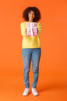 Full-length vertical shot cute friendly african-american woman, best friend stretching hands and giving lovely pink wrapped gift to you, smiling, congratulating with holidays, orange background.