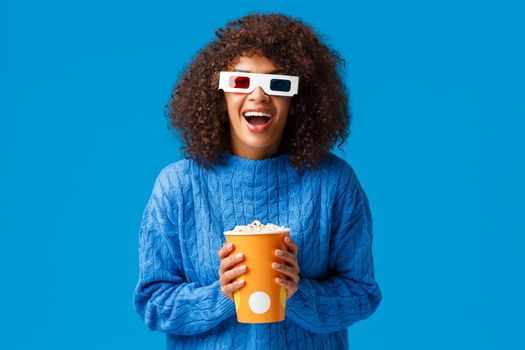 Cheerful gorgeous african-american woman enjoying watching movie in cinema, laughing and smiling excited with 3d effects wear glasses, holding popcorn, standing blue background.