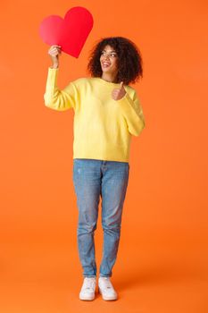 Full-length vertical shot satisfied and pleased, good-looking african american hipster girl feeling good without relationship, holding her red heart sign and give thumb-up, orange background.