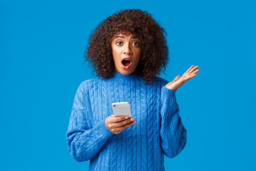Concerned and worried shocked young african-american woman receive unpleasant news via smartphone, telling it with indecisive look, shrugging and raise hand dont know what do, blue background.