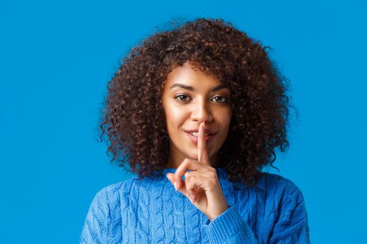 Close-up portrait sensual and mysterious african-american woman with cute happy smile, shushing, press index finger to lips having secret, prepare surprise and hush looking camera, blue background.