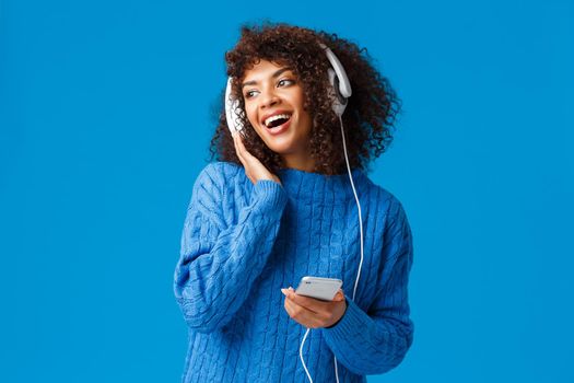 Charismatic modern young attractive african-american girl with afro haircut, listening music in headphones looking out window snowy weather, holding smartphone, smiling and singing.