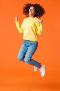 Full-length vertical shot happy and satisfied, carefree joyful african-american woman with afro, curly haircut, jumping from joy and happiness, showing thumbs-up over orange background.