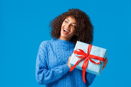 Curious and excited happy young african american woman shaking present to guess whats inside, standing intrigued receiving gift for christmas holidays, standing blue background.