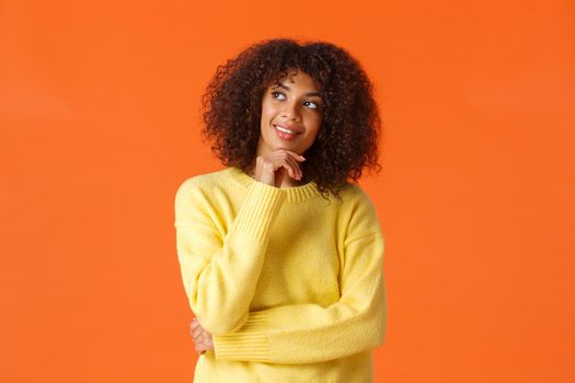 Pleased thoughtful, creative african american female entrepreneur, designer have interesting idea, smiling as thinking, planning something really good, know what present buy, orange background.