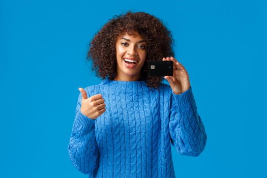 Satisfied young modern girl got bank account, opened deposit, using cashback service. Attractive african american woman in winter sweater, show thumbs-up and credit card in approval, recommend.