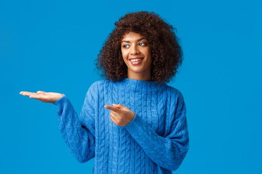 Charismatic lovely smiling african-american female helping family arrange table for holidays gathering, pointing finger left, presenting something on hand, standing blue background joyful.
