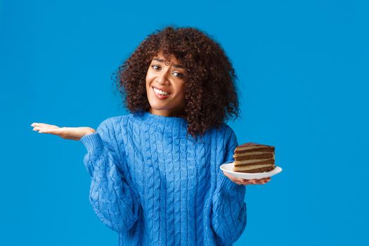 Unsure and tempting good-looking african-american girl taking care own body, looking after calories on diet, shrugging and raise hand clueless, holding plate with delicious cake, blue background.