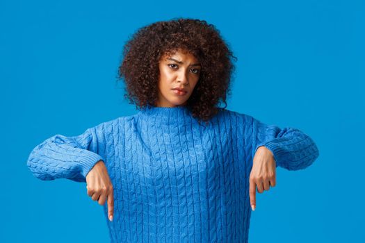 Upset and gloomy girl waiting for explanation, feeling uneasy and depressed, pointing fingers down, looking sad and unhappy camera, standing blue background distressed. Copy space