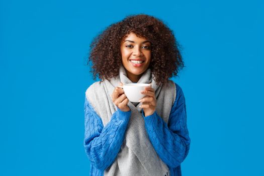 Girl feeling better after comming home and brewing delicious cup tea. Attractive smiling african-american happy woman with curly hair, wrap herself with scarf and drinking coffee, blue background.