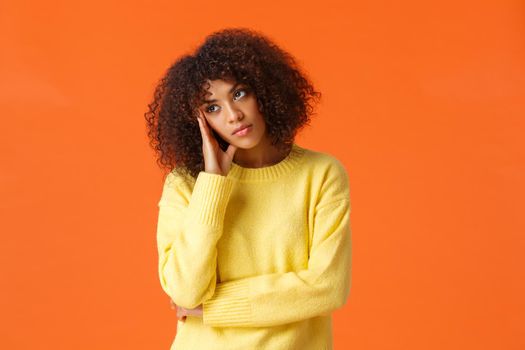 Uninterested bored african-american woman in yellow sweater, facepalm, looking away with uninterested, skeptical expression, attend boring party, standing upset and unsatisfied over orange background.