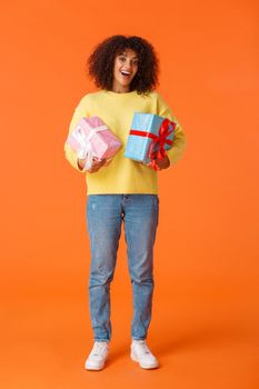 Full-length vertical shot excited cute happy african-american female received gifts for holidays, standing amused and delighted, holding two wrapped presents, orange background.