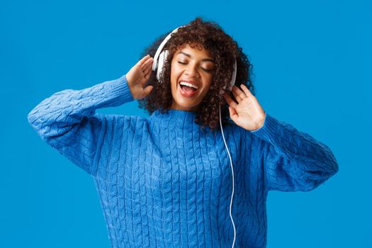 Waist-up portrait carefree happy smiling woman relaxing, singing along, playing karaoke app in earphones, close eyes passiontly lip-sync to music in headphones, standing blue background.