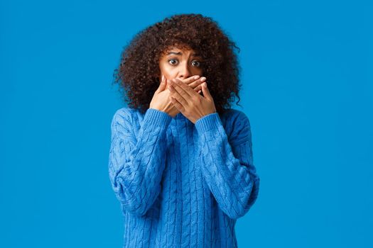 Scared and timid, insecure anxious young african-american woman witness terrible scene, gasping hold hands over mouth not scream, staring in panic and nervously standing over blue background.