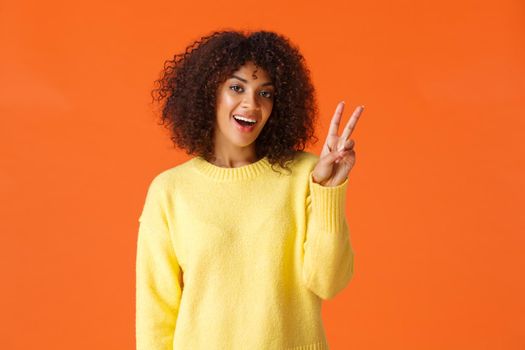 Carefree lovely, silly african-american girl posing for photo over orange background with smile, showing peace sign saying cheese feeling joyful and upbeat, express positive emotions.