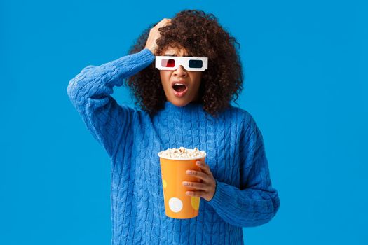 Shocked african-american pretty woman concerned with shocking cliffhanger in movie, grab head uneasy and upset, drop jaw, holding popcorn watching film in cinema, blue background.