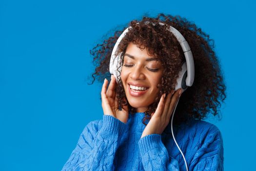 Close-up portrait tender and carefree happy smiling, sensual african-american woman in big headphones, close eyes and grinning romantic recall nice memories from listening song, blue background.