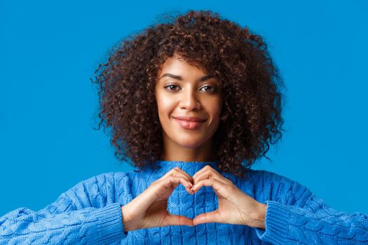 Close-up portrait tender and cute african-american romantic woman express her feelings with gesture, showing heart sign and smiling, have sympathy, confess over blue background.