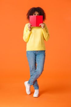 Full-length vertical shot shy and cute african-american modern urban girl with afro haircut, hiding face behind red notebook and peeking camera, writing in diary, standing orange background.