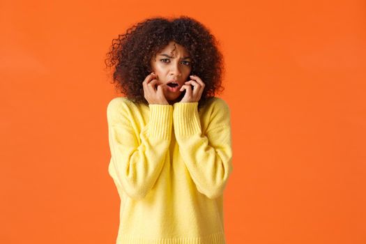 Waist-up portrait timid and insecure, scared cute african-american girl screaming and looking afraid at camera, holding hands pressed to face frightened, opened mouth gasping, orange wall.