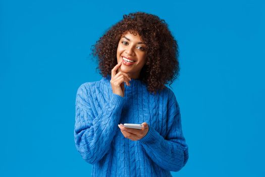 Hmm interesting. Intrigued and dreamy, creative african-american girl think-up christmas congratulation message, holding smartphone smiling and touching lip thoughtful, pondering, blue background.