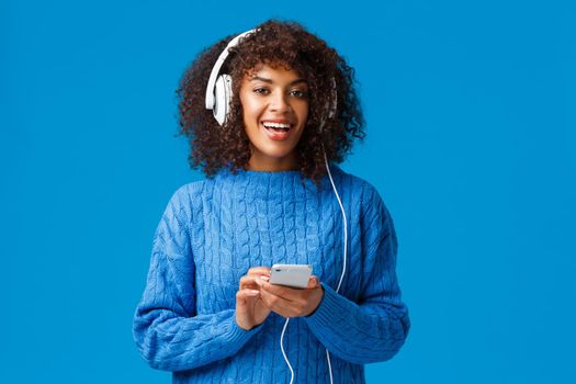 Carefree happy african-american hipster girl with afro haircut, put on headphones, turn-on favorite track and enjoying awesome sound quality in earphones, listen music, holding smartphone.