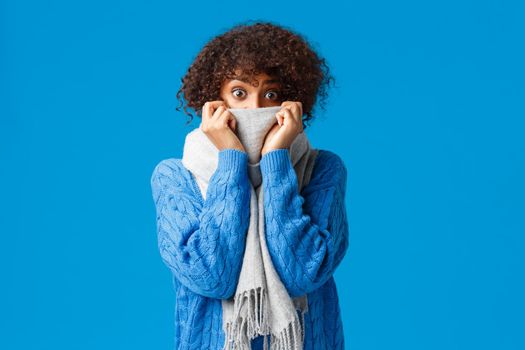 Silly scared and flushed cute african-american woman with afro haircut, hiding face behind scarf, staring popped eyes camera surprised or slightly anxious, standing blue background in winter sweater.