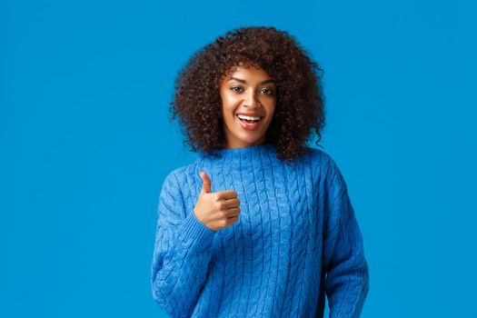 Nice choice, approve. Cute pretty african-american woman with afro haircut in winter sweater, showing thumb-up supportive, encourage friend did good job, satisfied standing blue background.