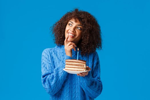 Dreamy and happy, beautiful african-american woman with afro haircut, thoughtful gazing up, smiling and touch lip as thinking what wish for before blowing-out candle in birthday cake.
