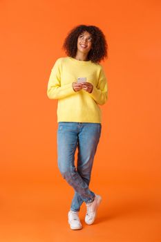 Full-length vertical shot thoughtful, lovely and dreamy african american woman with curly hair, looking up and smiling, imaging things, standing pensive, thinking what answer as holding smartphone.