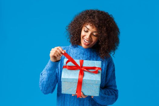 Curious and happy, smiling african-american woman, b-day girl in winter sweater, pulling present knot to unwrap gift and see what inside, celebrating christmas, new year holidays, blue background.