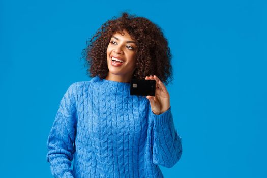 Cheerful attractive african-american woman with afro haircut, winter sweater, talking to friend looking left, smiling pleased, showing credit card, paying without cash, standing blue backgroun.