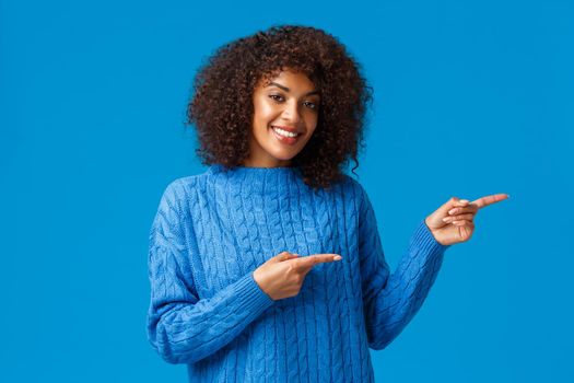 Lovely adult african-american female with afro haircut, pointing fingers right, suggesting go direction, visit site or download app, presenting great product and smiling satisfied, blue background.